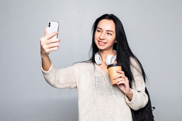 Retrato de una mujer atractiva sonriente tomando un selfie mientras sostiene llevar una taza de café aislada sobre la pared blanca