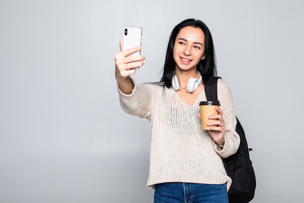Retrato de una mujer atractiva sonriente tomando un selfie mientras sostiene llevar una taza de café aislada sobre la pared blanca