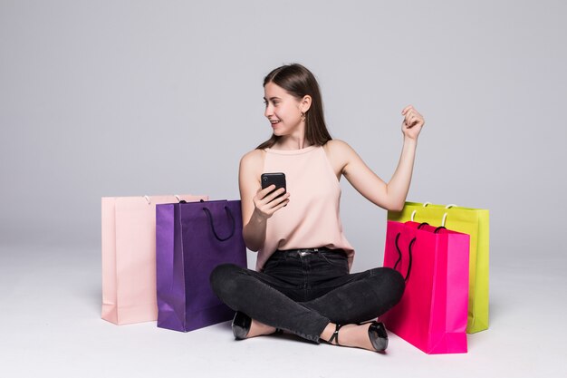 Retrato de una mujer bastante joven sentada en un piso con bolsas de compras y utilizando el teléfono móvil con gesto de ganar sobre la pared gris