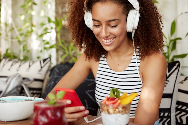 Retrato de mujer bloguera escucha canciones de audio en auriculares modernos, usa un teléfono inteligente para instalar una nueva aplicación, usa Internet gratis mientras toma una copa en la cafetería con un sabroso batido y un cóctel