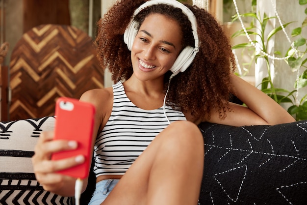 Foto gratuita retrato de mujer bloguera feliz publica nuevas fotos en el sitio web, hace autofotos en un teléfono inteligente moderno, escucha su música favorita en auriculares, pasa tiempo libre en un ambiente acogedor, usa wifi gratuito