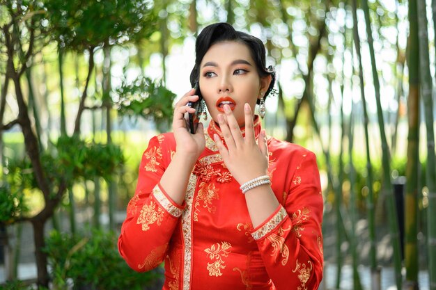 Retrato de mujer bonita asiática en un cheongsam chino posando emocionado durante hablar con el teléfono inteligente en el bosque de bambú