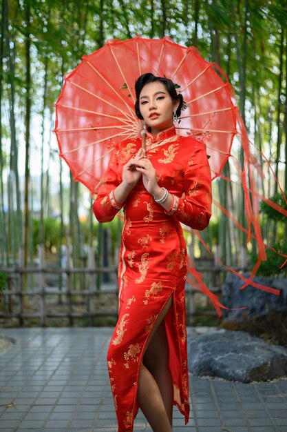 Retrato de mujer bonita asiática en un cheongsam chino posando con un hermoso paraguas de papel rojo, sonriendo y mirando a la cámara en el bosque de bambú, espacio de copia