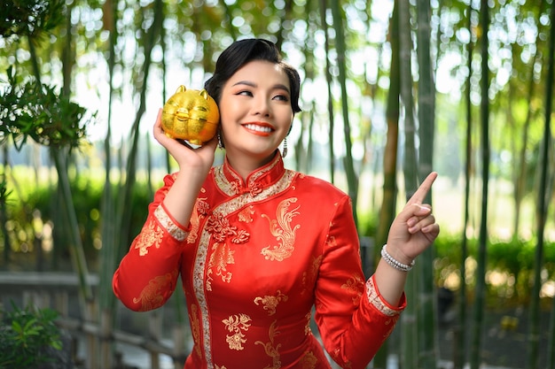 Retrato de mujer bonita asiática en un cheongsam chino posando con hucha y señalar con el dedo en el bosque de bambú