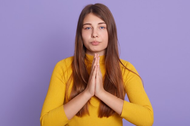 Retrato de mujer bonita con cabello largo y hermoso en camisa amarilla casual manteniendo las palmas juntas y rezando