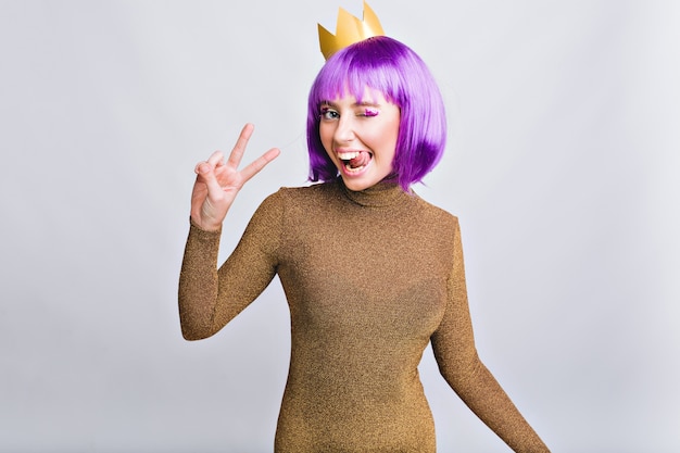 Retrato de mujer bonita con corona de oro divirtiéndose. Lleva un corte de pelo violeta, muestra la lengua y se ve feliz