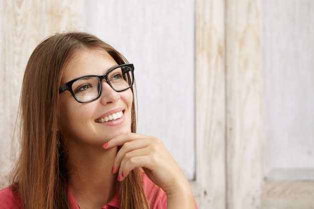 Retrato de mujer bonita en elegantes gafas rectangulares con expresión pensativa, sonriendo