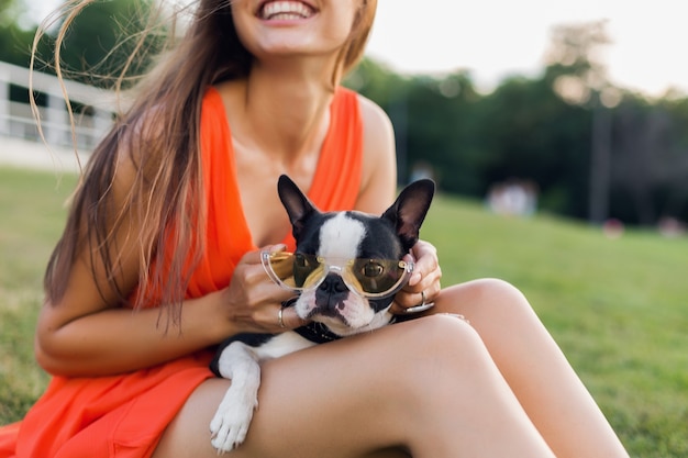 Retrato de mujer bonita feliz sentada en el césped en el parque de verano, sosteniendo el perro boston terrier, humor positivo sonriente, vestido naranja, estilo moderno, gafas de sol, jugando con mascota