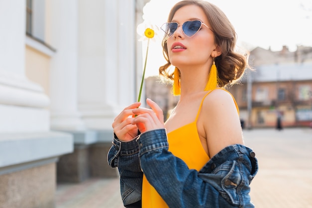 Retrato de mujer bonita con gafas de sol de corazón con flor contra el sol, día soleado de verano, ropa elegante, tendencia de moda, chaqueta de jeans azul, vestido amarillo, elegantes pendientes boho hipster