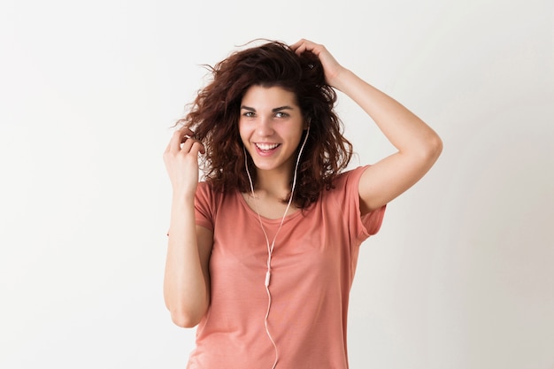 Retrato de mujer bonita hipster feliz sonriente de aspecto natural joven con peinado rizado en camisa rosa posando aislado sobre fondo blanco de estudio, escuchando música en auriculares