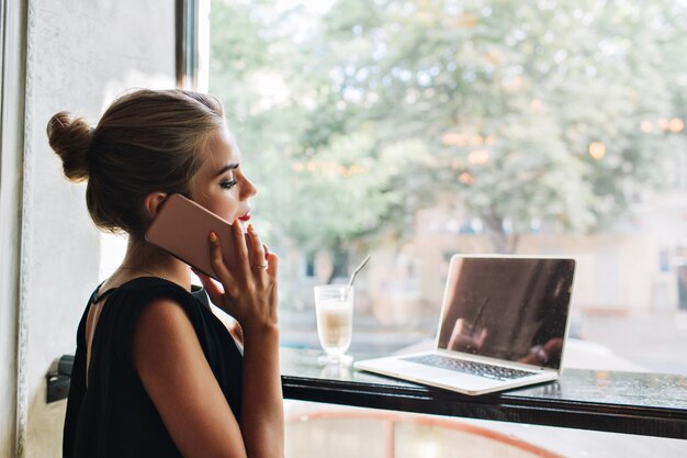 Retrato de mujer bonita de lado en vestido negro en la cafetería. Ella está hablando por teléfono, mirando en la computadora portátil.