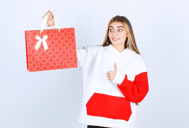 Retrato de mujer bonita modelo de pie con bolsa de regalo y mostrando el pulgar hacia arriba