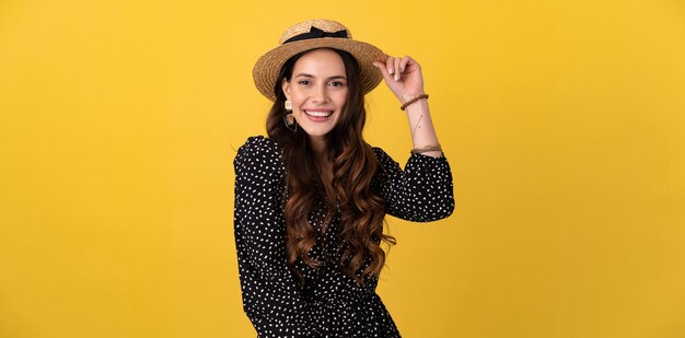 Retrato de mujer bonita posando aislado en amarillo con vestido negro punteado y sombrero de paja con estilo boho tendencia