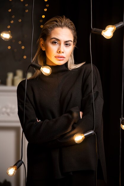 Retrato de mujer bonita posando en suéter oscuro de pie junto a las lámparas.