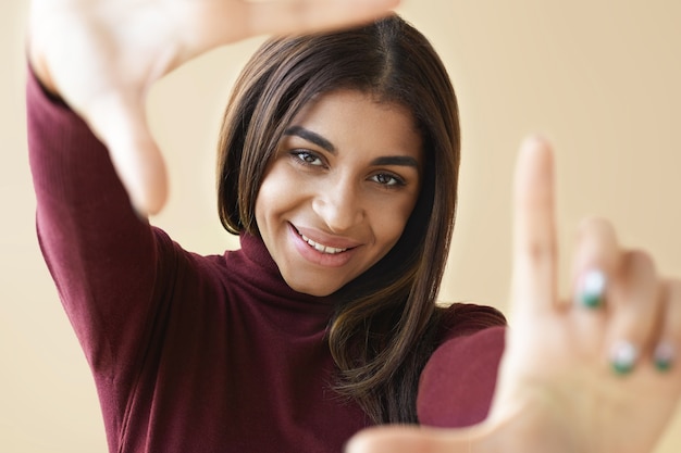 Retrato de mujer bonita de raza mixta de cerca sonriendo felizmente y gesticulando, haciendo marco con sus dedos, mirando a través de él como si estuviera tomando una foto. Enfoque selectivo en la cara de la niña.