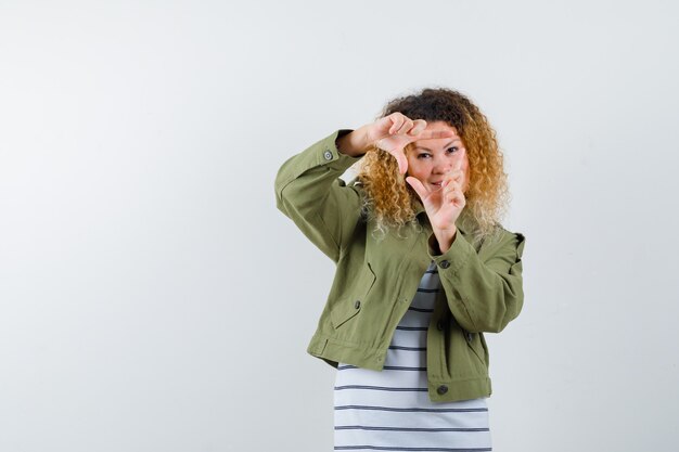 Retrato de mujer bonita rubia haciendo gesto de marco en chaqueta verde y mirando confiada vista frontal