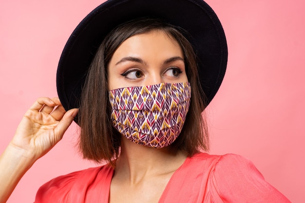 Retrato de mujer bonita vestida con máscara protectora elegante de cerca. El uso de sombrero negro y gafas de sol. Posando sobre pared rosa