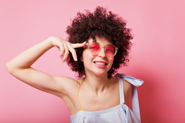 Retrato de mujer brillante con rizos cortos viste gafas rosas y camisa azul en rosa