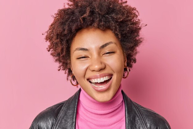 El retrato de una mujer de buen aspecto con una amplia sonrisa dentuda expresa emociones felices, se siente alegre, muestra dientes blancos, lleva cuello de tortuga y chaqueta de cuero aislada sobre fondo rosa. concepto de felicidad