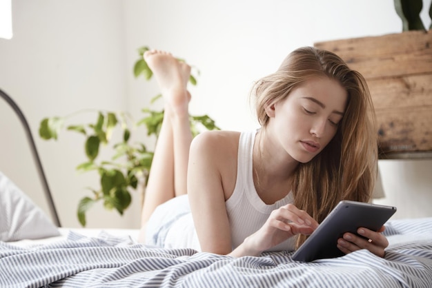 Retrato de mujer buscando en internet en tableta yace en la cama por la mañana