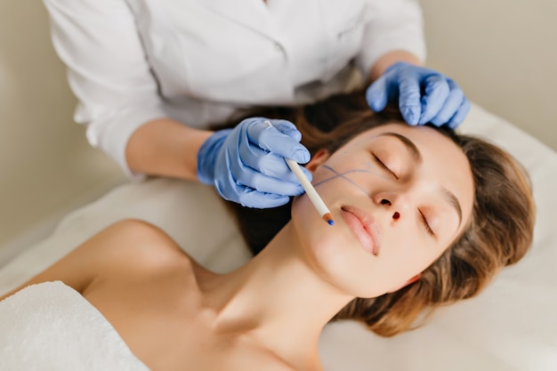 Retrato de mujer con cabello morena en preparación para el rejuvenecimiento, operación de cosmetología en salón de belleza. Manos en guantes azules dibujando en la cara, botox, belleza