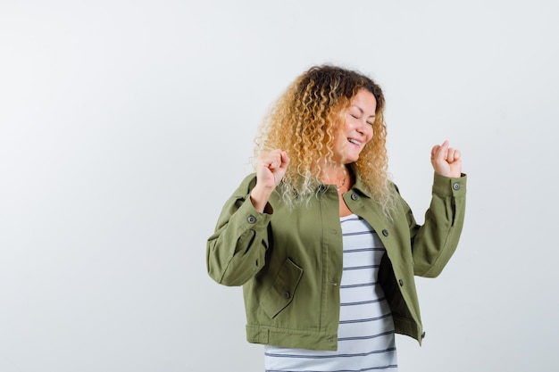 Retrato de mujer con cabello rubio rizado mostrando gesto ganador en chaqueta verde y mirando feliz vista frontal