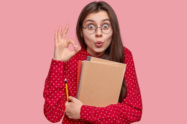 Retrato de mujer con camisa roja y anteojos redondos sosteniendo blocs de notas