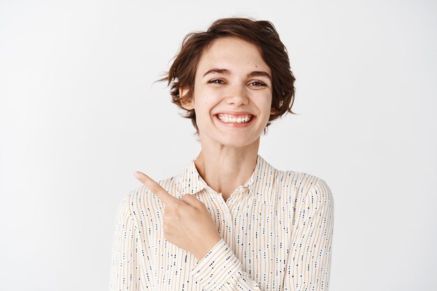 Retrato de mujer caucásica en blusa sonriendo y apuntando con el dedo en la esquina superior izquierda, de pie sobre la pared blanca