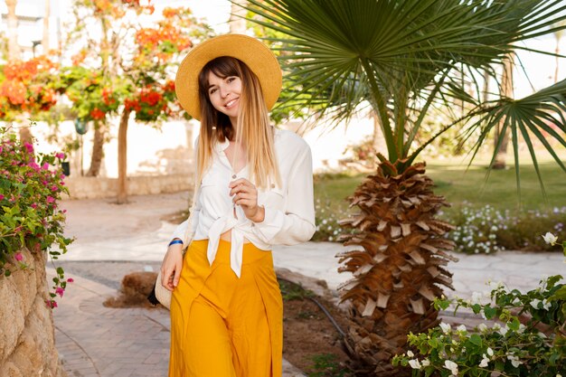 Retrato de mujer caucásica bonita en sombrero de paja, blusa blanca y bolso de estilo Bali caminando en jardín tropical.