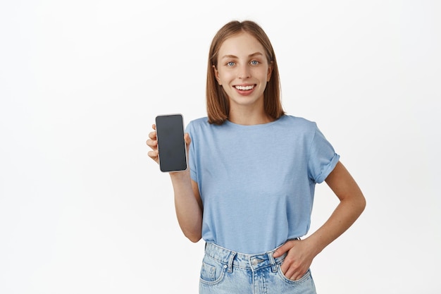 Retrato de mujer caucásica feliz que muestra la pantalla del teléfono móvil, sonriendo satisfecha, recomendando la aplicación, buena interfaz de teléfono inteligente, de pie contra el fondo blanco