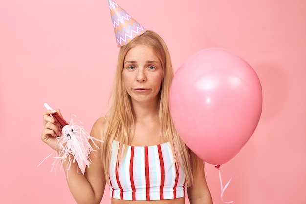 Foto gratuita retrato de mujer caucásica joven con estilo molesto con top rayado y sombrero cónico sosteniendo globo de helio después de haber decepcionado la expresión facial aburrida de estar triste y solo