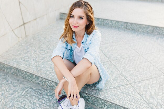 Retrato de una mujer caucásica joven feliz que se sienta en los pasos grises claros que sonríen.