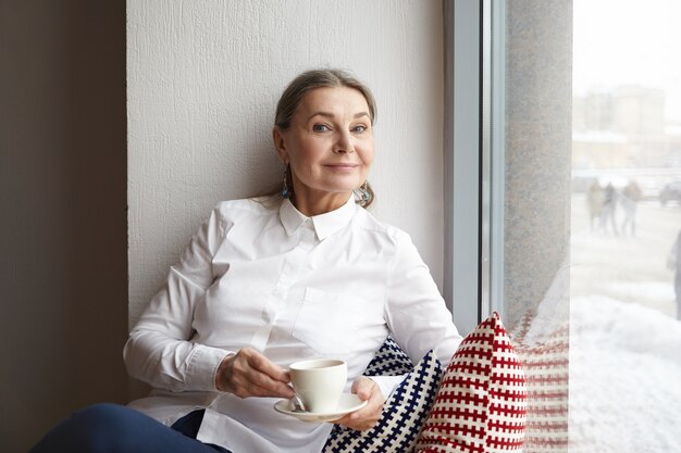 Retrato de mujer caucásica madura elegante atractiva en camisa blanca relajante en la cafetería con taza de capuchino, sentado en el alféizar de la ventana y sonriendo felizmente. Concepto de personas y estilo de vida