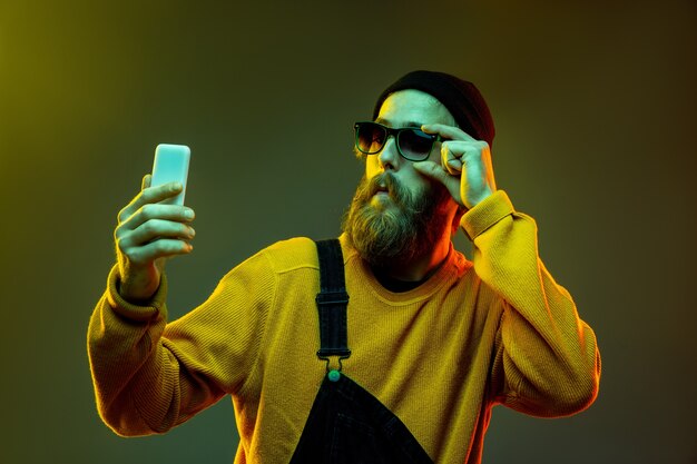 Retrato de mujer caucásica sobre fondo de estudio degradado en luz de neón. Hermoso modelo masculino con estilo hipster en gafas. Concepto de emociones humanas, expresión facial, ventas, publicidad. Usando el teléfono.