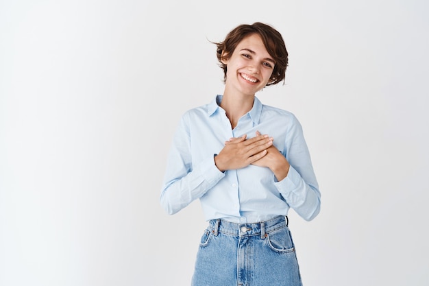 Retrato de mujer caucásica sonriente tomados de la mano en el corazón y agradeciéndole, sintiéndose agradecido, de pie en la pared blanca