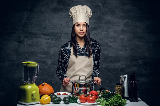 Retrato de mujer chef con una olla en la mesa con verduras frescas.