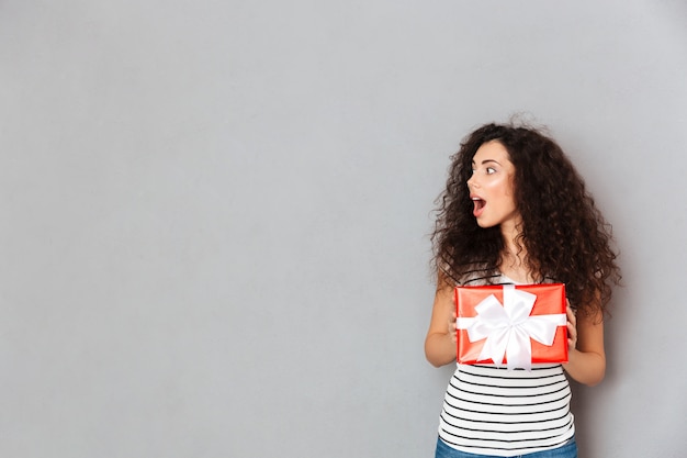 Foto gratuita retrato de mujer contenta y sincera con cabello oscuro y rizado con caja roja envuelta para regalo, emocionada y sorprendida copia espacio