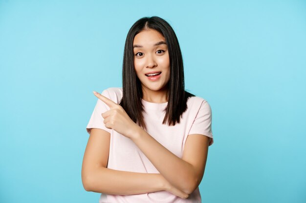 Retrato de mujer coreana sonriente apuntando a la esquina superior izquierda, mostrando descuento, banner de venta, de pie en camiseta sobre fondo azul.