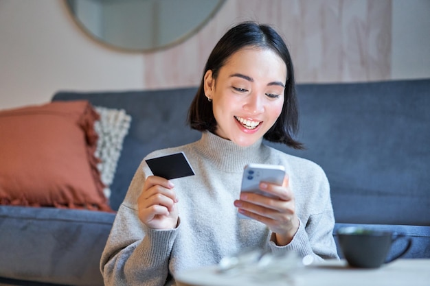 Foto gratuita retrato de mujer coreana sonriente usando tarjeta de crédito y aplicación de teléfono inteligente pagando la orden de compra onli