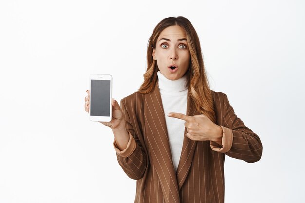 Retrato de una mujer corporativa apuntando a un teléfono móvil con una expresión de asombro que muestra el sitio web en la interfaz de la aplicación para smartphone con fondo blanco