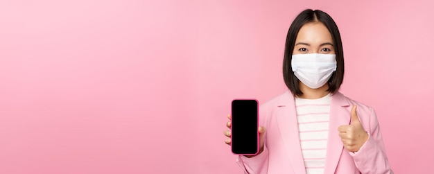 Retrato de una mujer corporativa asiática con mascarilla médica covid19 que muestra la pantalla del teléfono inteligente y el pulgar arriba muestra el fondo rosa