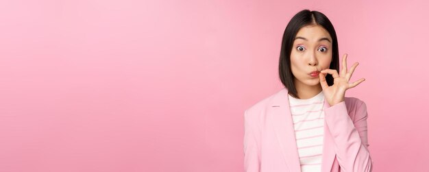 Retrato de una mujer corporativa asiática que muestra el sello de la boca cerrar los labios cerrados en gesto clave promete mantener el secreto de pie sobre fondo rosa en traje