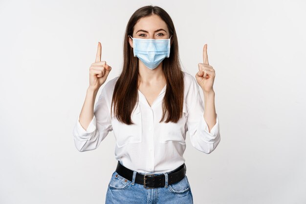 Retrato de mujer corporativa con máscara médica facial de coronavirus señalando con el dedo hacia arriba mostrando un anuncio...