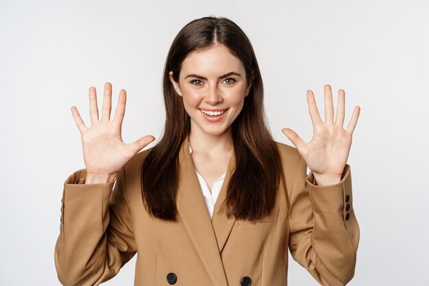 Retrato de mujer corporativa, vendedora mostrando el número diez dedos y sonriendo, de pie en traje sobre fondo blanco.