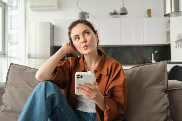 Foto gratuita retrato de una mujer curiosa y linda sentada en el sofá con un teléfono inteligente mira hacia arriba y piensa en colocar un