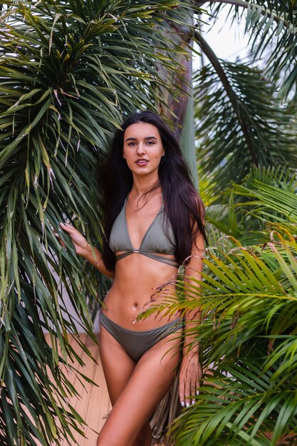 Retrato de mujer delgada bronceada en forma en bikini pequeño verde posando con hojas tropicales