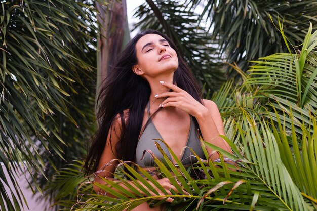 Retrato de mujer delgada bronceada en forma en bikini pequeño verde posando con hojas tropicales