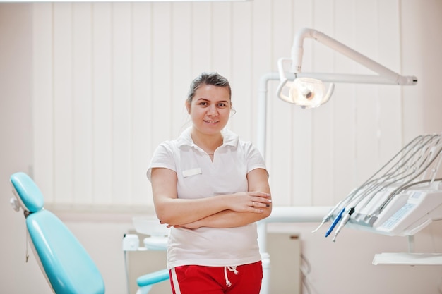 Retrato de mujer dentista cruzó los brazos de pie en su oficina de odontología cerca de la silla