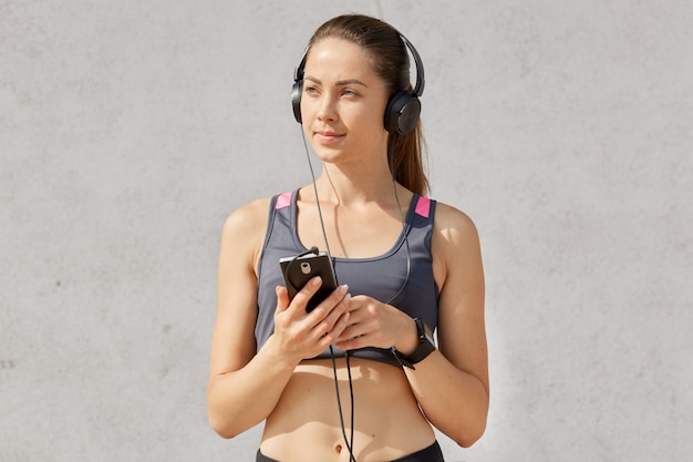 Foto gratuita retrato de mujer deportiva atractiva en sujetador deportivo escuchando música con auriculares y teléfono inteligente, tiene cola de caballo