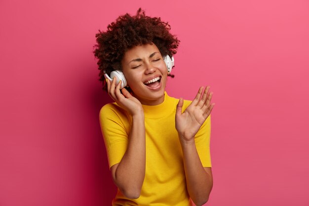 Retrato de mujer despreocupada llena de alegría escucha música, canta canciones usa auriculares, cierra los ojos, se olvida de todos los problemas, usa ropa amarilla, aislada en la pared rosa. Concepto de estilo de vida